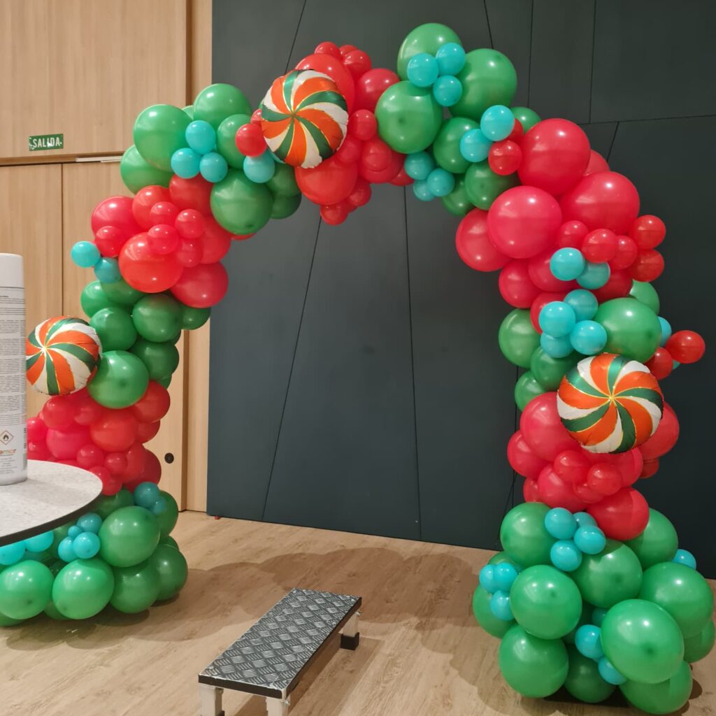 Colorido arco de globos en tonos rojos, verdes y azul claro, decorado con caramelos gigantes de globos, ideal para fiestas temáticas y celebraciones infantiles.