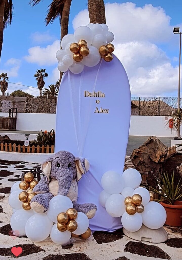 Decoración de fiesta infantil con globos blancos y dorados, cartel personalizado y oso de peluche en un entorno exterior con palmeras y cielo azul. Ideal para celebraciones temáticas y cumpleaños infantiles.